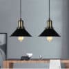 Agda Loft Black English Street Style Pendant Light - Two black, industrial-style pendant lights with cone-shaped shades hang over a wooden dining table. The lights create a warm and inviting atmosphere in the room.