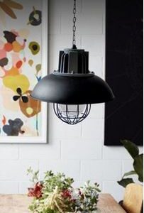 Alfhild Oval Industrial Pendant Light - A black industrial pendant light hanging over a wooden dining table. The light has a wide, black shade and a cage-like structure around the bulb. A colorful painting and a vase of flowers are visible in the background.