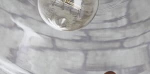 A person's hand installing a light fixture with a clear glass shade. The background shows a brick wall.